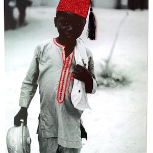 Op de afbeelding: Een zwart-witfoto van een persoon die een rode fez met een zwarte kwast draagt, een lichtgekleurde tuniek met rode borduursels en een waterkan draagt. De persoon houdt ook een witte doek vast.