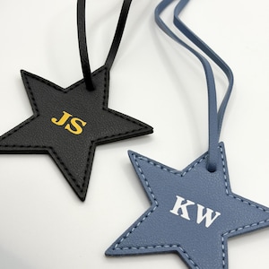 May include: Two star-shaped luggage tags. One is black with gold lettering "JS", the other is blue with white lettering "KW". Both have matching colored straps.