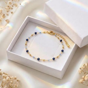 Könnte beinhalten: Ein zartes, goldfarbenes Kettenarmband mit abwechselnd dunkelblauen, kugelförmigen Perlen und kleineren hellblauen Perlen, ausgestattet mit einem Karabinerverschluss und einer Verlängerungskette, präsentiert in einer weißen, quadratischen Geschenkbox.