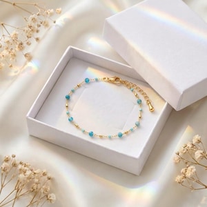 Könnte beinhalten: Ein zartes Armband aus einer goldfarbenen Kette mit kleinen, runden, leuchtend blauen Perlen, die gleichmäßig verteilt sind. Das Armband hat einen goldenen Karabinerverschluss und eine verstellbare Verlängerungskette mit einem kleinen tropfenförmigen Anhänger.