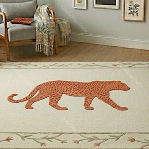 May include: A rectangular area rug featuring a cream-colored background with a central orange leopard motif. The rug is bordered by a delicate vine pattern with small orange flowers, creating a distinct frame around the central animal figure.