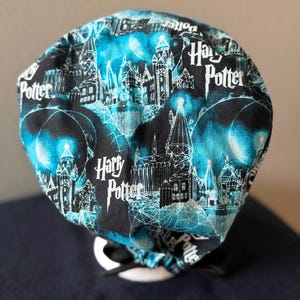 Può includere: Un cappello chirurgico nero e verde acqua con un motivo ripetuto del castello di Hogwarts, lune e le parole "Harry Potter" in bianco. Il cappello ha un fiocco nero ed è fatto di tessuto.