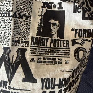 Può includere: Un oggetto in tessuto bianco con un design a stampa di giornale in bianco e nero. Il design presenta le parole "Harry Potter", un ritratto di Harry Potter e vari elementi di testo, che ricordano un manifesto di ricerca.
