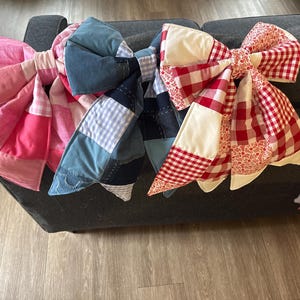 May include: Three large, decorative fabric bows in shades of pink, blue, and red. The bows are made from quilted patchwork fabric, with gingham and floral patterns. The bows are arranged on a dark gray surface.