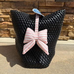 Può includere: Borsa tote nera intrecciata con un fiocco a righe rosa e bianche e un accento decorativo blu. La borsa ha una trama intrecciata e una forma rettangolare. Il fiocco è centrato sulla parte anteriore della borsa.