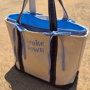 May include: A cream-coloured canvas tote bag with black handles and a black base. The bag features a bright blue interior lining and a blue and white gingham trim with tied bows on the sides. The text "broke down" is embroidered in blue.