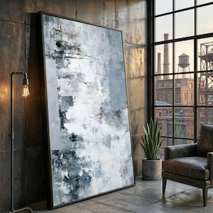 May include: Large framed abstract artwork with a textured design in white, grey, and black. The painting leans against a metal wall in an industrial setting. A floor lamp and a leather armchair are also visible in the room.