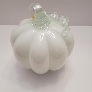 May include: A decorative glass pumpkin featuring a smooth, opaque white body with deep vertical ridges. The top of the pumpkin is adorned with a clear, ribbed glass stem that curves and twists into a vine-like shape, adding texture to the piece.