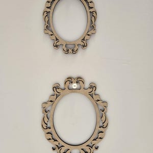 May include: Two oval-shaped frames made of light brown wood with intricate, symmetrical scrollwork patterns along the edges. Each frame features a small, white, circular bead or pin centered at the top of the ornate border design.