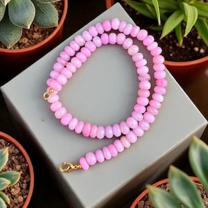 May include: A pink beaded necklace with a gold clasp. The necklace features small, rounded beads in shades of pink. The necklace is displayed on a grey surface, with potted plants in the background.