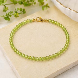 May include: A delicate bracelet featuring small, faceted, translucent lime-green peridot beads strung in a circular pattern. The bracelet is secured with a polished gold-tone metal lobster claw clasp and matching gold-tone metal hardware components.