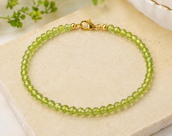 May include: A delicate bracelet featuring small, faceted, translucent lime-green peridot beads strung in a circular pattern. The bracelet is secured with a polished gold-tone metal lobster claw clasp and matching gold-tone metal hardware components.