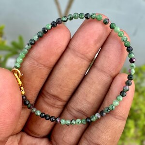 May include: A beaded bracelet featuring small, faceted, round gemstone beads in shades of green, black, and occasional flecks of deep pink. The bracelet is secured with a gold-toned metal lobster claw clasp and a small jump ring connector.