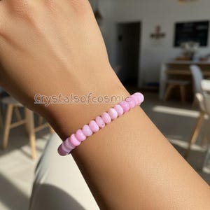 Peut inclure: Un bracelet de perles rose clair est porté sur un bras. Le bracelet est composé de petites perles rondes. Le texte "Crystalsofcosmic" est visible sur le bras. Le bracelet est un bijou délicat.