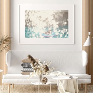 May include: A framed art print depicting a blurred, sun-dappled pool scene. The artwork hangs above a white sofa with decorative pillows and a vase of dried flowers. Two small, white side tables and a floor lamp complete the interior design.