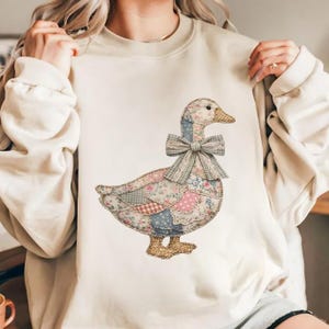 May include: Cream-colored sweatshirt featuring a patchwork goose design. The goose has a floral patterned body, a blue and floral neck, and a gray bow. The design is centered on the front of the sweatshirt, offering a cozy and whimsical aesthetic.