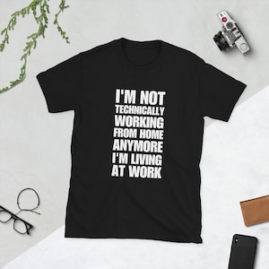 Könnte beinhalten: Ein schwarzes T-Shirt mit kurzen Ärmeln und weißem Text auf der Vorderseite, der lautet: I'M NOT TECHNICALLY WORKING FROM HOME ANYMORE I'M LIVING AT WORK. Das T-Shirt hat einen Rundhalsausschnitt und besteht aus einfarbigem schwarzem Stoff.