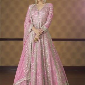 May include: A pink, floor-length gown with intricate silver embroidery. The dress features a V-neckline, long sleeves, and a flowing skirt. The gown is adorned with detailed patterns and embellishments, creating a luxurious and elegant look.