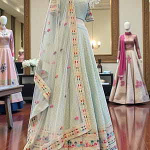 May include: A light green lehenga, a traditional Indian outfit, displayed on a mannequin. The skirt and short-sleeved top are embellished with floral embroidery and a matching sheer dupatta. The outfit is displayed in a boutique setting.