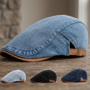 May include: A blue denim flat cap featuring a structured crown, a curved brim, and a tan suede-like adjustable side strap with a metallic buckle. The design includes visible stitching along the panels and a smooth, rounded silhouette.