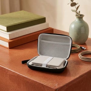May include: An open black zippered case with a white interior, holding a small, rectangular device secured by a white strap. The case sits on a reddish-orange surface, with books and a vase in the background. Cables and a small green vase are also visible.