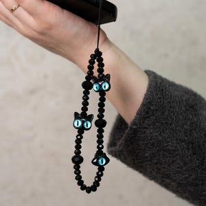 May include: Black beaded phone charm featuring three cat-shaped charms with blue eyes. The charm is attached to a black phone. The beads are various shapes and sizes, creating a unique accessory.
