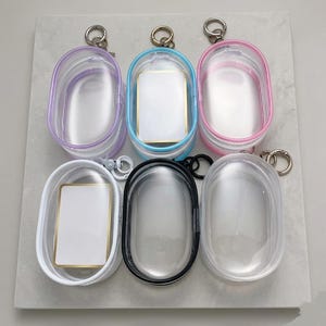 Puede incluir: Seis estuches ovalados transparentes con ribetes de colores y llaveros plateados. Los estuches tienen ribetes morados, azules, rosas, blancos, negros y transparentes. Cada estuche tiene un cierre de cremallera y un pequeño inserto rectangular.