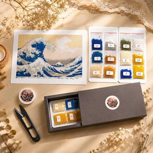 Op de afbeelding: Een diamond painting set met een bedrukt canvas van De Grote Golf bij Kanagawa, twaalf genummerde zakjes met gekleurde kralen, een donkergrijze opbergdoos met extra kralenzakjes, een rond bakje en een zwarte metalen schaar.