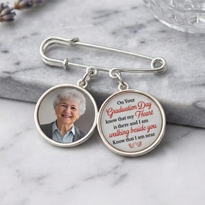 May include: A silver-colored graduation pin with two circular pendants. One pendant features a photo of a smiling woman. The other pendant has the text "On Your Graduation Day know that my Heart is there and I am walking beside you."