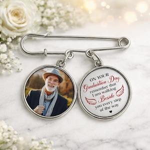 May include: A silver-tone memorial pin with two circular pendants. One pendant features a photo of a smiling man wearing a hat and scarf. The other pendant has the text "ON YOUR Graduation Day, remember that I am walking Beside you every step of the way."