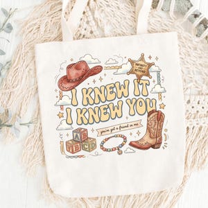 May include: A cream-colored canvas tote bag featuring a graphic print with a brown cowboy hat, a sheriff star badge labeled Infinity and Beyond, alphabet blocks, a beaded bracelet, and a cowboy boot with the text I KNEW IT I KNEW YOU.