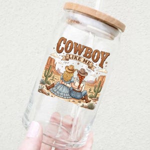 Pode incluir: Um copo de vidro transparente com tampa de bambu e palha, apresentando um gr&aacute;fico colorido de duas pessoas com chap&eacute;us de cowboy sentadas numa paisagem des&eacute;rtica com cactos, com o texto COWBOY LIKE ME impresso acima da ilustra&ccedil;&atilde;o.
