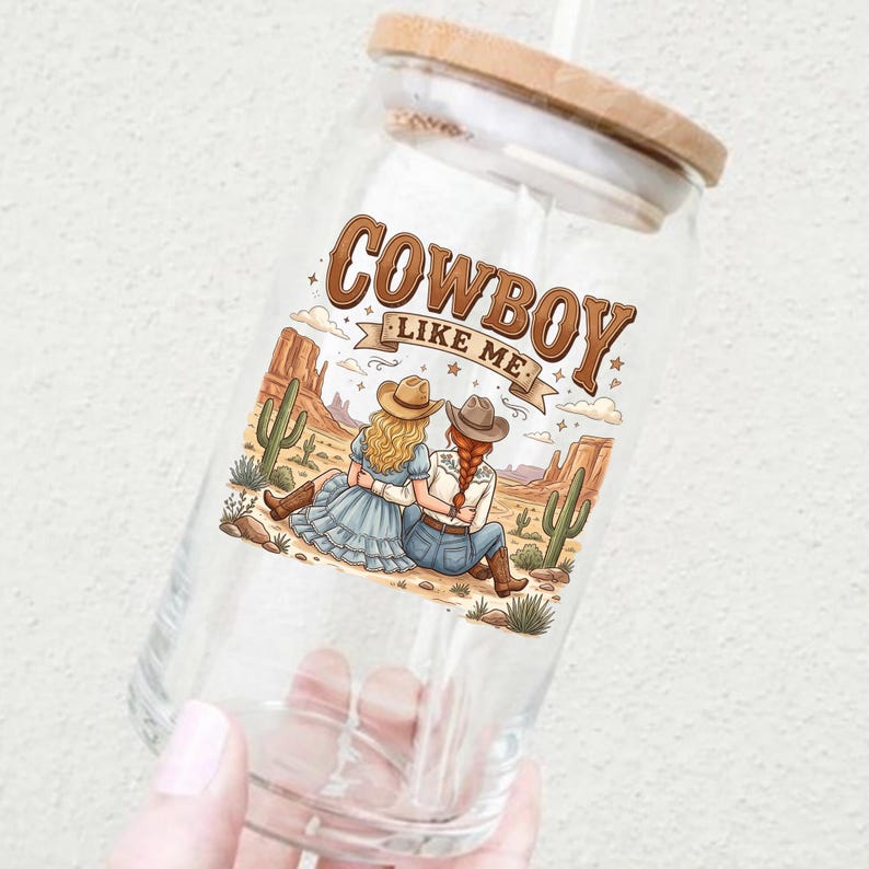 Pode incluir: Um copo de vidro transparente com tampa de bambu e palha, apresentando um gr&aacute;fico colorido de duas pessoas com chap&eacute;us de cowboy sentadas numa paisagem des&eacute;rtica com cactos, com o texto COWBOY LIKE ME impresso acima da ilustra&ccedil;&atilde;o.