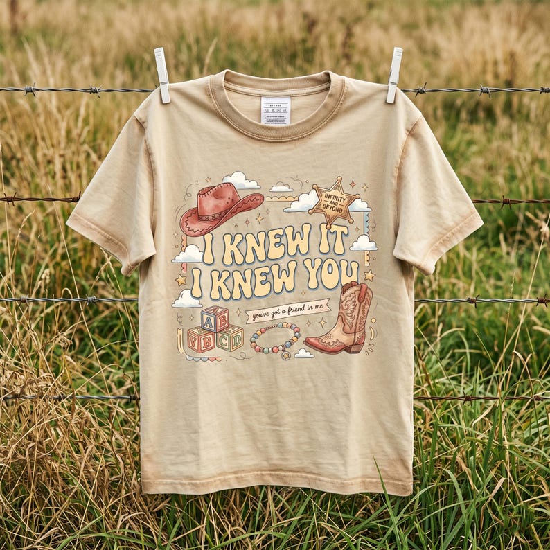 May include: A beige short-sleeve t-shirt featuring a graphic print with a cowboy hat, a sheriff badge labeled "INFINITY AND BEYOND," alphabet blocks, a beaded bracelet, and a cowboy boot, with the text "I KNEW IT I KNEW YOU" and "you've got a friend in me."