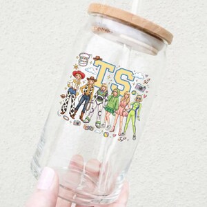 K&ouml;nnte beinhalten: Ein durchsichtiges Glas mit Bambusdeckel und Strohhalm, das mit einem bunten Grafikdruck von sechs stilisierten Figuren in verschiedenen Outfits, darunter ein Raumanzug und Cowgirl-Kleidung, sowie den gro&szlig;en gelben und blauen Buchstaben TS versehen ist.