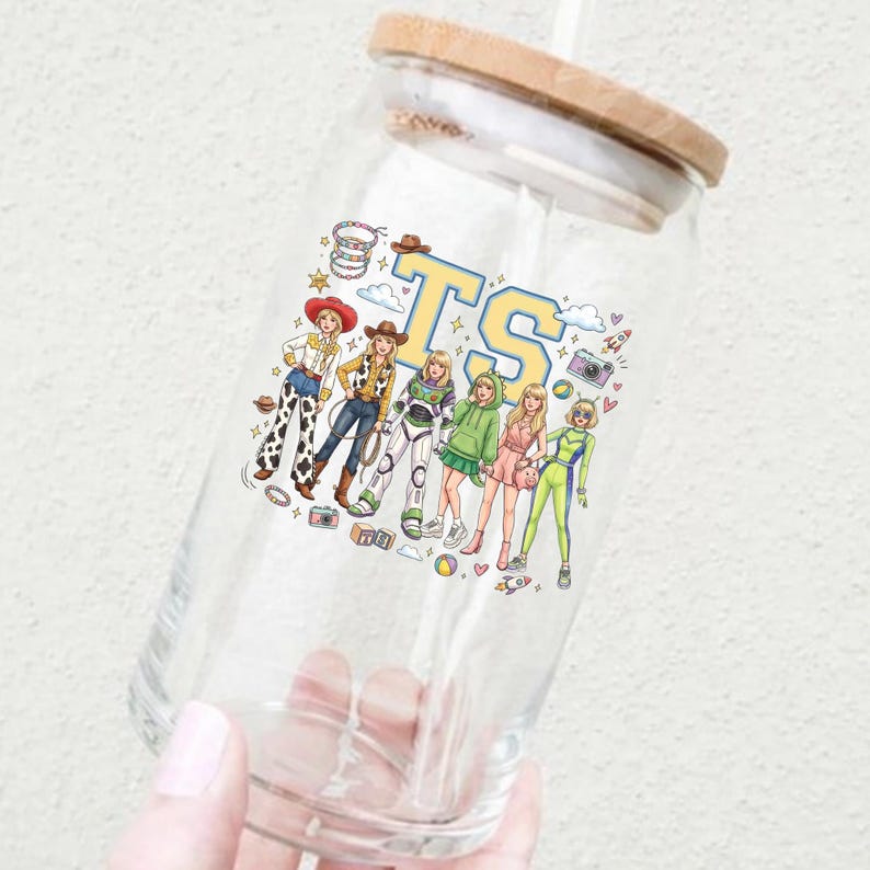 K&ouml;nnte beinhalten: Ein durchsichtiges Glas mit Bambusdeckel und Strohhalm, das mit einem bunten Grafikdruck von sechs stilisierten Figuren in verschiedenen Outfits, darunter ein Raumanzug und Cowgirl-Kleidung, sowie den gro&szlig;en gelben und blauen Buchstaben TS versehen ist.