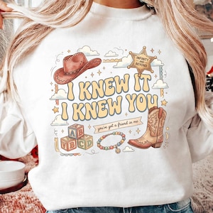 May include: A white sweatshirt featuring a graphic print with a brown cowboy hat, a sheriff star badge labeled Infinity and Beyond, alphabet blocks, a beaded bracelet, and a cowboy boot. The text reads I KNEW IT I KNEW YOU and you've got a friend in me.
