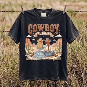 Pode incluir: Uma t-shirt preta de manga curta com uma estampa gr&aacute;fica de duas pessoas usando chap&eacute;us de cowboy e botas, sentadas numa paisagem des&eacute;rtica com cactos. O texto "COWBOY LIKE ME" est&aacute; impresso numa fonte castanha grande e estilizada.