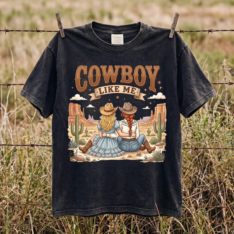 Pode incluir: Uma t-shirt preta de manga curta com uma estampa gr&aacute;fica de duas pessoas usando chap&eacute;us de cowboy e botas, sentadas numa paisagem des&eacute;rtica com cactos. O texto "COWBOY LIKE ME" est&aacute; impresso numa fonte castanha grande e estilizada.