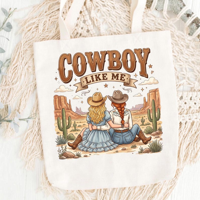 Pode incluir: Uma sacola de lona cor de creme com uma estampa gr&aacute;fica de duas pessoas usando chap&eacute;us de cowboy sentadas em uma paisagem des&eacute;rtica com cactos e forma&ccedil;&otilde;es rochosas. O texto "COWBOY LIKE ME" est&aacute; impresso em letras marrons acima da ilustra&ccedil;&atilde;o.