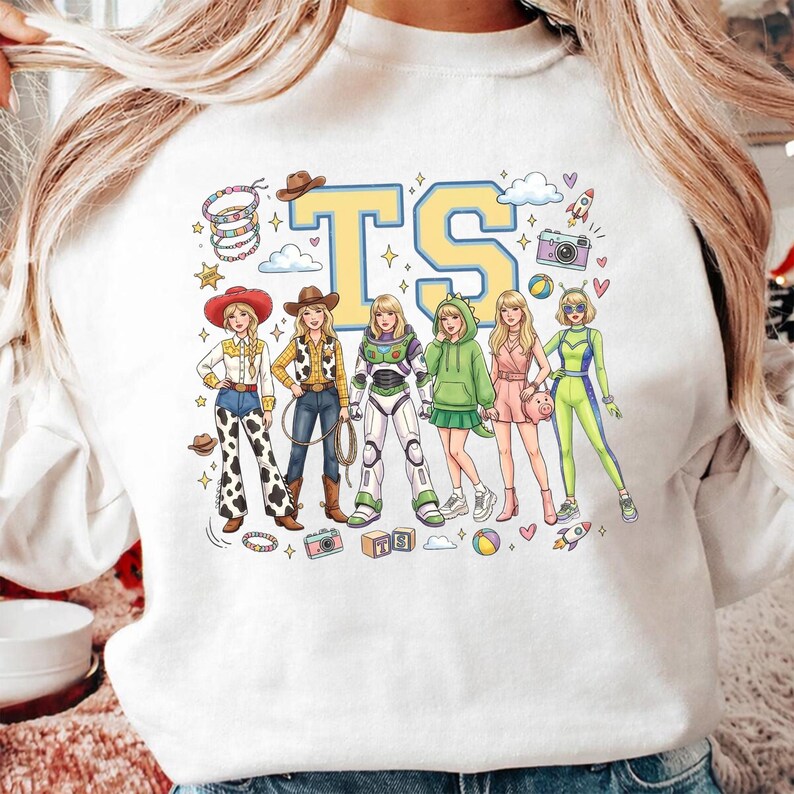 K&ouml;nnte beinhalten: Ein wei&szlig;es Sweatshirt mit einem Grafikdruck von sechs illustrierten Figuren in verschiedenen Outfits, darunter Cowboy-Kleidung, ein Weltraum-Ranger-Anzug und ein gr&uuml;ner Dinosaurier-Kapuzenpullover. Die Buchstaben TS sind in gro&szlig;en gelben Blockbuchstaben dar&uuml;ber gedruckt.