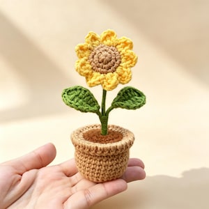 Puede incluir: Un girasol tejido a mano en un tiesto pequeño. El girasol tiene pétalos amarillos, un centro marrón, hojas verdes y un tallo verde. El tiesto es de color marrón claro.