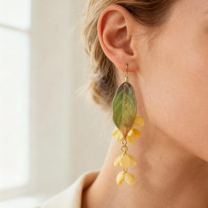 Op de afbeelding: Goudkleurige oorhangers met een bladvorm en gele bloemaccenten. De oorbellen hebben een haaksluiting en zijn gemaakt van metaal en hars. Het blad is groen en bruin, en de bloemen zijn zachtgeel.