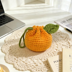 Pode incluir: Uma bolsa laranja de crochê, em forma de laranja, com um caule e uma folha verdes. A bolsa com cordão está sobre uma toalha de crochê creme. Um laptop e livros estão no fundo.