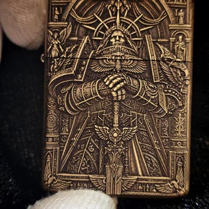 Puede incluir: Un encendedor rectangular de metal color latón con un grabado intrincado en relieve profundo de una figura central blindada con una espada, rodeada de motivos alados, arcos arquitectónicos y figuras pequeñas.