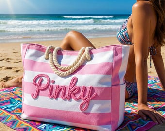 May include: A large pink and white striped tote bag with the word "Pinky" in pink script. The bag has rope handles and is sitting on a colorful beach towel. The background shows the ocean and a sunny sky.