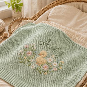 May include: A light green knitted baby blanket with an embroidered duck and floral design. The name "Avery" is embroidered above the duck. The blanket is placed in a wicker bassinet with a cream-colored lining.