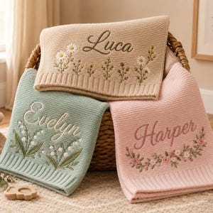 Può includere: Tre coperte lavorate a maglia in beige, verde salvia e rosa tenue, ognuna con disegni floreali ricamati e nomi personalizzati. La coperta beige mostra il nome Luca, quella verde Evelyn e la coperta rosa presenta il nome Harper.