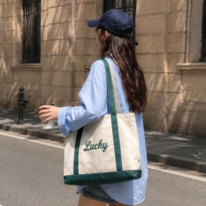 May include: A cream-coloured canvas tote bag featuring dark green trim along the bottom and handles. The word Lucky is embroidered in dark green cursive script on the front of the bag. The bag has two long shoulder straps and a rectangular shape.