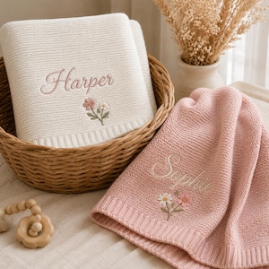 Puede incluir: Una manta de punto color crema con el nombre Harper bordado y un pequeño motivo floral, junto a una manta de punto rosa con el nombre Sophie bordado y un diseño floral a juego, ambas dentro de una cesta de mimbre tejida.